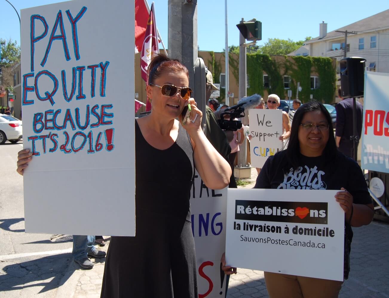 Deux femmes pancarte à la main : « Pay Equity because it’s 2016 » et « Rétablissons la livraison à domicile ».